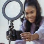 Cheerful young African American female blogger in stylish sweater smiling while setting up camera of smartphone attached to tripod with ring light before recording vlog