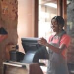 Cheerful American African waitress in apron working on counter monitor while registering order at cozy cafe