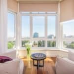 Interior of modern penthouse with comfortable white armchairs and round side table placed near windows overlooking city in sunny day