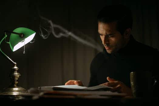 Pondering young male in black sweater reading book while sitting at desk with papers and green bankers lamp in dark home office