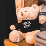 Small teddy bear in black t shirt with inscription FUCK OFF placed on wooden shelf in shop