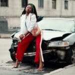 Woman in White Open Cardigan and Red and White Pants Sitting on Damage White Car