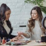 Young positive multiethnic female bloggers testing makeup products while sitting at table and recording vlog on smartphone placed on tripod with ring light