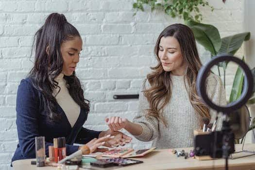 Young positive multiethnic female bloggers testing makeup products while sitting at table and recording vlog on smartphone placed on tripod with ring light