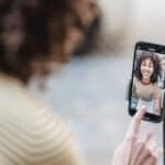 Crop cheerful young ethnic female vlogger touching screen on cellphone while preparing for video record in town