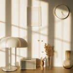 Light room with retro radio and decorative vases with dry plants on desk near wall with clock and window shadow in sunlight