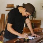 Side view of Asian man in casual clothes and eyeglasses bending on table and taking notes by pencil while working in joinery