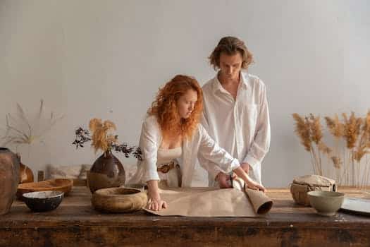 Concentrated man and redhead woman standing near wooden table with craft paper placed near bowl during packaging in cozy workshop