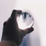 Crop anonymous person demonstrating transparent glass ball in hand against white wall with upside down reflection of modern tall skyscrapers in daylight