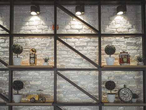 Different clocks with ornament and potted plants on wooden shelves near rough wall illuminated by shiny lamps in house