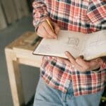 From above of crop anonymous slim male carpenter in plaid shirt and blue jeans doing quick sketches of future furniture in notepad while leaning on workbench in workshop