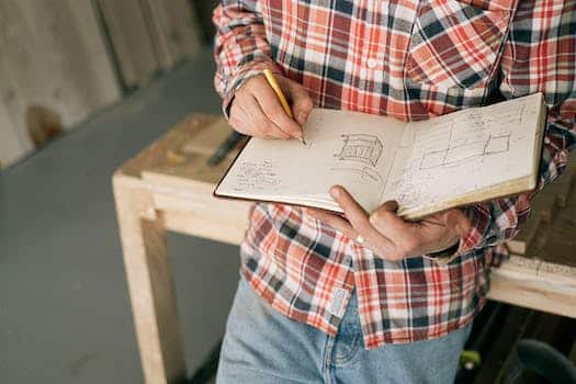 From above of crop anonymous slim male carpenter in plaid shirt and blue jeans doing quick sketches of future furniture in notepad while leaning on workbench in workshop