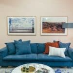 Big comfortable blue sofa with soft cushions placed in modern living room with round marble table near wall decorated with framed pictures