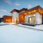 Exterior architecture of private suburban cottage house with stone and wooden facade and large windows overlooking spacious snow covered yard in winter day