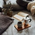 From above of cup of hot coffee with pot on wooden tray near open book and dry plant in cozy bed